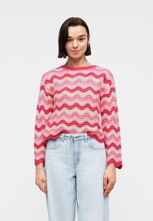 Joven con cabello corto y oscuro, vistiendo un suéter de patrones ondulados en rosa y crema y jeans de tiro alto de color azul claro sobre un fondo blanco.