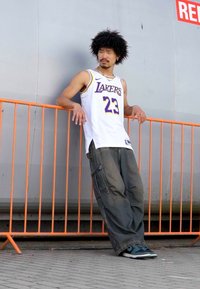 Bijela košarkaška dres s ljubičastim natpisom "Lakers" i brojem 23, uparen s labavim sivim cargo hlačama i plavim tenisicama. Narančasta barijera u pozadini.