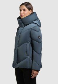 Giacca puffy di colore blu scuro, con colletto alto, design trapuntato a rombi e logo sulla manica. Presenta tasche laterali e una texture liscia.