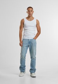 Jaunas vyras stovi prieš paprastą foną, vilkėdamas baltą marškinėlį be rankovių, šviesiai mėlynus džinsus ir baltus sportbačius, su viena ranka kišenėje.