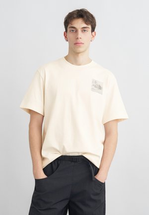 Jeune homme portant un t-shirt crème The North Face et un pantalon noir, debout les mains dans les poches devant un fond uni.