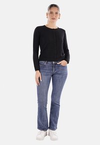 Cardigan nero a maniche lunghe con bottoni frontali, abbinato a jeans blu a zampa d'elefante e sneakers bianche. Texture liscia e design aderente.