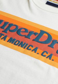 Hvid t-shirt med stort blåt "Superdry" tekst, prydet med horisontale striber i orange og røde nuancer. Glat stoftekstur og crew neck design.