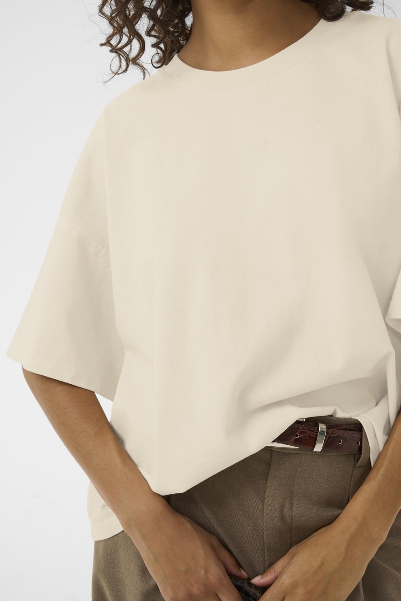 Übergroßes cremefarbenes T-Shirt mit kurzen Ärmeln, Rundhalsausschnitt und dezentem Branding. Kombiniert mit beigen Hosen und einem braunen Gürtel.