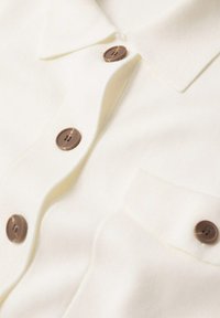Chemise blanche à col avec des boutons marron, dotée d'une poche avant et de poignets boutonnés sur un tissu doux.
