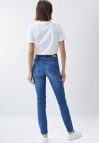 Jeans ajustados en denim azul medio, con una cintura alta, bolsillos traseros y costuras sutiles. Combinados con una camiseta blanca básica y zapatillas blancas.