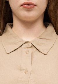 Κοντινό πλάνο από το κάτω μέρος προσώπου και λαιμό ενός ατόμου που φοράει μπεζ πουκάμισο με γιακά, κουμπωμένο στην κορυφή.