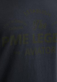 Camiseta oscura de color navy con texto grande y en negrita en verde olivo. El texto incluye "PME LEGEND" y "AVIATOR" con acentos decorativos. Material de algodón suave.