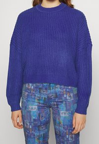 Avskuren lila stickad tröja med en texturerad väv och långa ärmar. Kombineras med blå mönstrade byxor som har abstrakta mönster i olika nyanser.