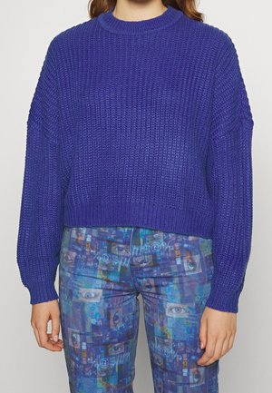 Pull en maille violet coupé avec un tissage texturé et des manches longues. Porté avec un pantalon bleu à motifs comportant des designs abstraits dans des nuances variées.