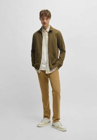Olive green zip-up jacket over a light plaid shirt, paired with tan pants and white sneakers. Smooth material, casual design, and fitted style.