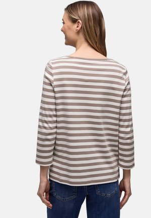 Vrouw die een beige-wit gestreept shirt met lange mouwen en blauwe spijkerbroek draagt, met haar rug naar de camera toe, tegen een effen achtergrond.