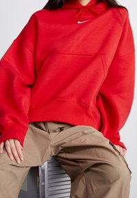 Röd hoodie med en framficka, snörning i huvan och liten vit logga. Mjuk material och oversized passform, kombinerad med beige baggy byxor.