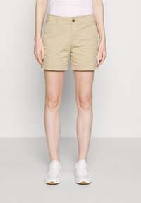 Shorts kaki en tissu mélangé de coton lisse, avec une fermeture à boutons sur le devant et des revers roulés, associés à des baskets blanches.