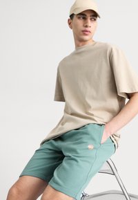 Camiseta beige con cuello redondo, combinada con pantalones cortos de algodón verdes que tienen un pequeño logo. Modelo sentado en una silla de metal.