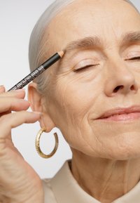 Une personne tient un crayon à sourcils noir, l'appliquant sur son sourcil. Elle porte de grandes boucles d'oreilles en or et un haut à col de couleur claire.