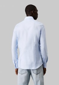 Lichtblauw, langwerpig shirt met een gestructureerd patroon, knopenmanchetten en een traditionele kraag, gezien vanuit de achterkant.
