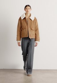Bruine wolmix jas met een witte schapenvacht kraag, voorzien van een rits aan de voorkant en twee grote zakken. Gecombineerd met grijze broek en sneakers.