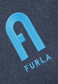 Modře vyšívané stylizované písmeno "F" a text "FURLA" na tmavě texturovaném pozadí.