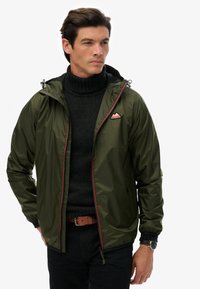 Olivgrön vattentät jacka med dragkedja fram, justerbar huva och röda detaljer. Bärs över en svart ribbad polotröja. Svarta jeans.