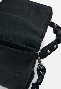 Sac à bandoulière en cuir noir avec surface texturée, fermeture éclair, sangle réglable et accents en métal argenté. Il dispose d'une petite poche avant.