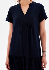 Kvinna i en kortärmad marinblå klänning med V-ringning och kedjehalsband, som visar armarna med en synlig orm-tatuering på höger underarm.