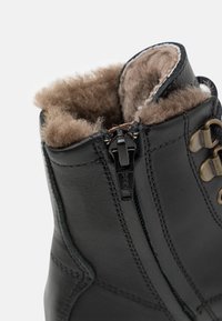 Bota de cuero negro con un forro marrón suave, cremallera frontal y herrajes de acento metálico. El diseño incluye detalles cosidos en la superficie.