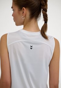 Vitt ärmlöst träningsshirt med en texturerad yta, med en svart chevron-logotyp på baksidan nära halsringningen.