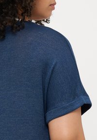 Μπλε πλεκτό μπλουζάκι με κοντά ρολαρισμένα μανίκια, που διαθέτει υφασμάτινη υφή και χαλαρή εφαρμογή. Ο λαιμός στο πίσω μέρος έχει ελαφρώς στρογγυλό σχήμα.