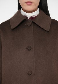 Cappotto in lana marrone con ampio colletto, chiusura frontale con quattro bottoni circolari e una texture morbida.