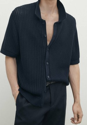 Mann trägt ein dunkelblaues, kurzärmeliges Strickhemd mit Knöpfen und strukturiertem Muster, kombiniert mit schwarzen Hosen, steht mit den Händen an den Seiten.