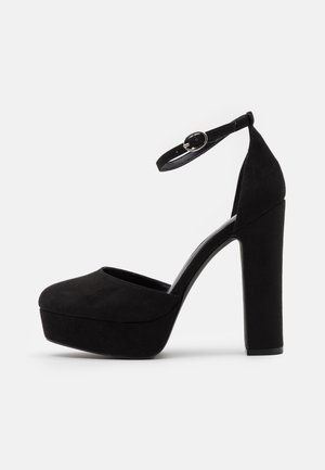 Black suede platform heels with a rounded toe, ankle strap, and a chunky high heel. Smooth texture with a solid black color.