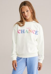 Jauna meitene ar gariem gaisvīriem brūniem matiem valkā baltu džemperi ar daudzkrāsainu tekstu "ŅEMIZIŅU" un gaiši zilas ribotas bikses.