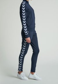 Navy träningsställ tillverkat av mjukt tyg, bestående av en zip-up jacka och tighta byxor med vita chevron-accenter längs sidorna. Vita sneakers.