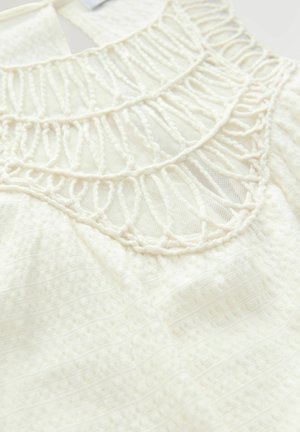 Haut en tissu blanc avec des détails en crochet complexe au niveau du col, texture douce et fond à motif avec des variations subtiles.