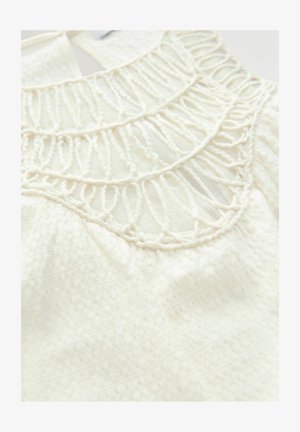 Biela tkanina s vrchnou časťou, ktorá sa vyznačuje zložitým háčkovaním na výstrihu, jemnou textúrou a vzorovaným pozadím s jemnými variáciami.