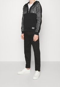 Svart hoodie med dragkedja, med ett mönstrat övre parti, framlomma och logotyp. Kombinerad med svarta byxor och vita sneakers.