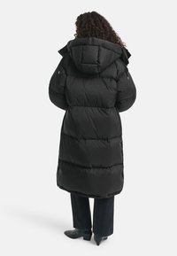 Persoon met krullend haar, gekleed in een lange zwarte gewatteerde jas met capuchon, zwarte broek en zwarte hakken, staand met de rug naar de kijker.