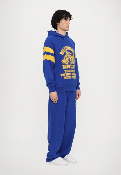 Blaue Kapuzenpullover und passende Jogginghose mit gelben Streifen und Logo-Print. Hergestellt aus weichem Stoff, mit lockerer Passform und Taschen.