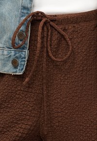 Κοντινό πλάνο καφέ υφασμάτινου παντελονιού με υφή, δεμένα στη μέση με κορδόνια, συνδυασμένα με κουμπιά ανοιχτό μπλε τζιν μπουφάν και λευκό ύφασμα από κάτω.