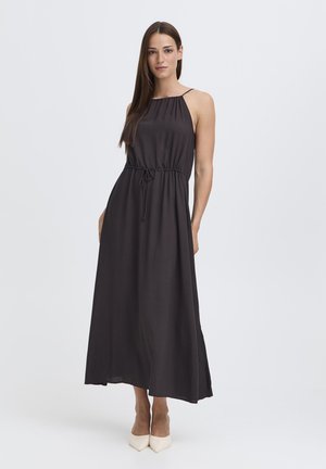 Moteris stovinti vilkinti juodą, be rankovių, ilgesnę suknelę su siaurinta liemeniu ir baltus aukštakulnius su smailiomis noselėmis, ant šviesaus paprasto fono.