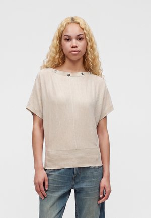 Ung kvinde med langt krøllet blondt hår iført en beige kortærmet top med knapper og blå jeans, der står foran en simpel baggrund.