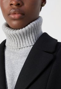Siv oblekast pulover s turtleneck ovratnikom pod črnim blejzerjem, s prilegajočim ovratnikom in mehko teksturo, ki poudarja slojevitost oblačil.