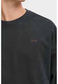 Svart hettegenser i bomull med ribbet rund hals og en liten, brodert logo på venstre bryst. Jevn tekstur.