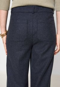 Vue arrière d'une personne portant un pantalon en laine gris foncé avec deux poches arrière plaquées et un pull en ribou beige, les mains sur les côtés avec des bracelets en or.