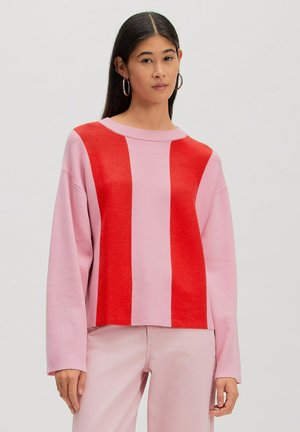 Frau mit glatten dunklen Haaren, die rosa Hosen und einen pink-roten vertikal gestreiften Pullover trägt, stehend vor einem schlichten Hintergrund.