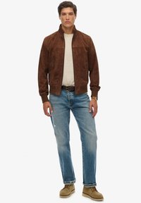 Giacca bomber in suede marrone con colletto alto, abbinata a una camicia beige chiaro, jeans in denim blu e scarpe beige. Design semplice, vestibilità casual.