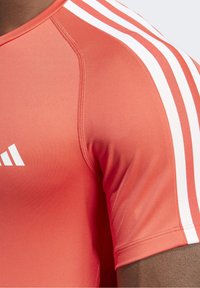 Camisa atlética roja con rayas blancas en los hombros. Tejido suave y elástico con corte ajustado. Presenta detalles de costura cerca del cuello.