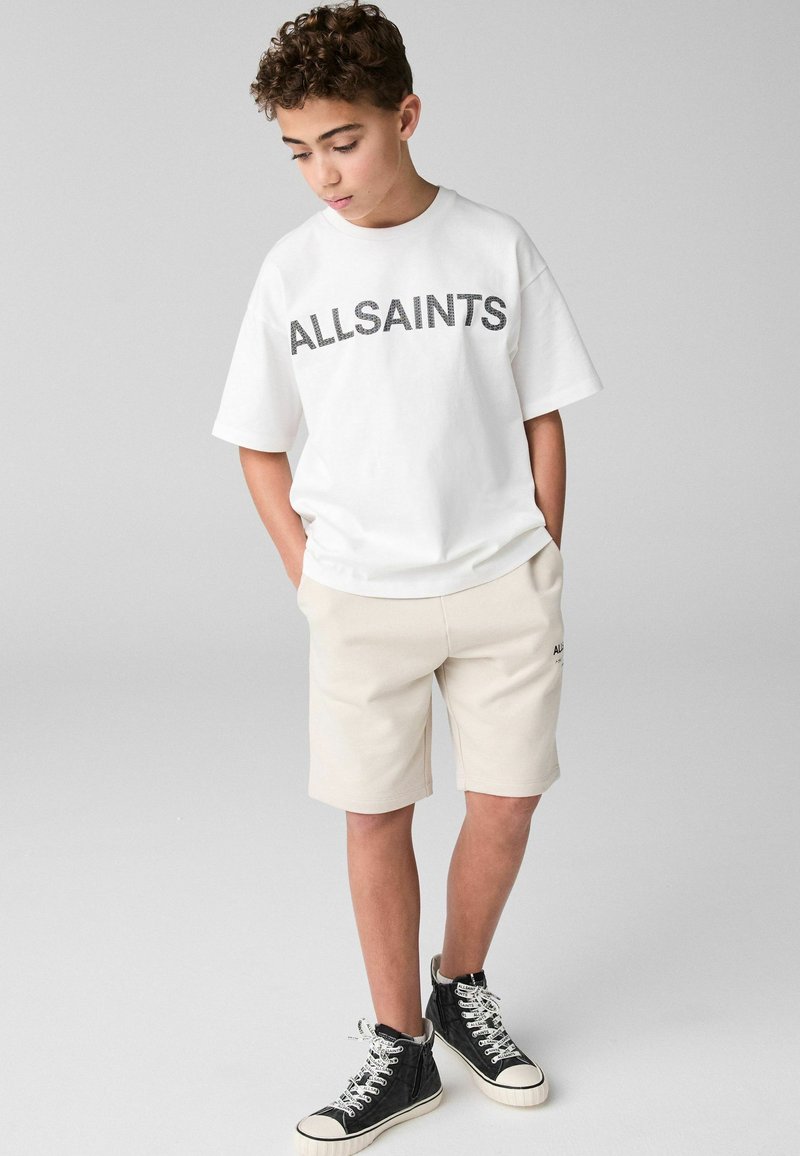 Vit bomull t-shirt med "ALLSAINTS" tryckt i svart, tillsammans med beiga shorts. Svarta höga sneakers med vita sulor.