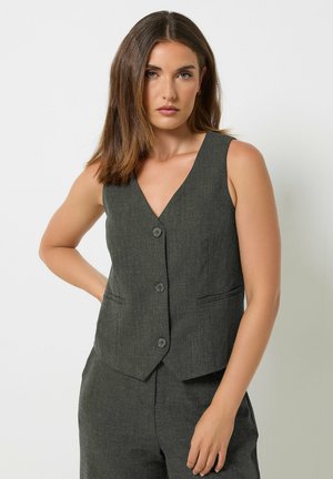 Woman with shoulder-length hair wearing a dark gray buttoned vest and matching pants, standing with one hand on her hip against a plain background.